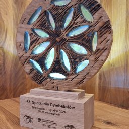 IKSD - Usługi Wycinania Grawerowania Laserowego - Drewniana nagroda z wygrawerowanym wzorem kwiatowym i datą wydarzenia, stojąca na drewnianej podstawie z napisem '43. Spotkania Cymbalistów'.