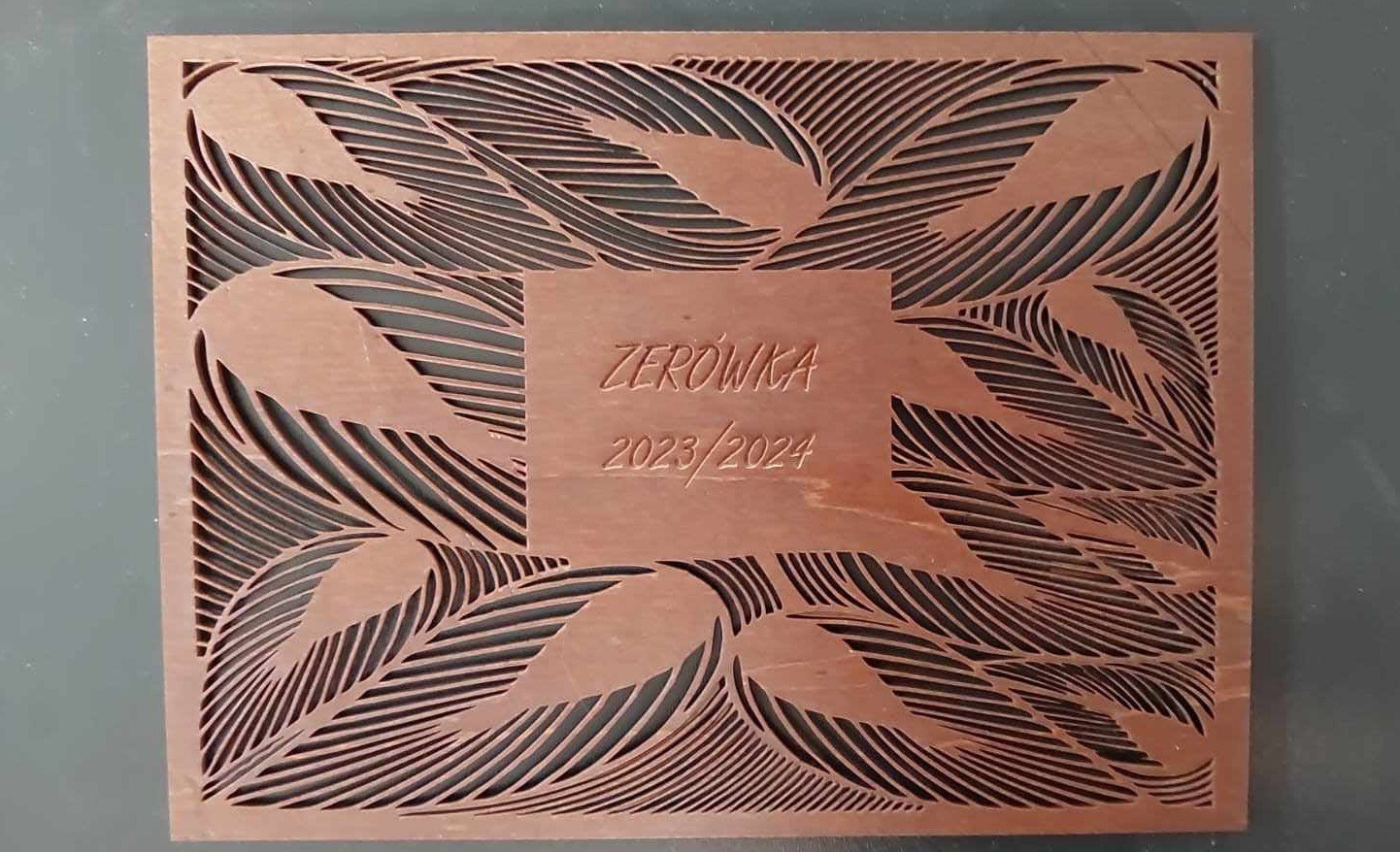 Drewniana tabliczka z wyciętym laserowo wzorem liści i napisem 'Zerówka 2023/2024' w centralnym kwadracie. Precyzyjne cięcie CNC.