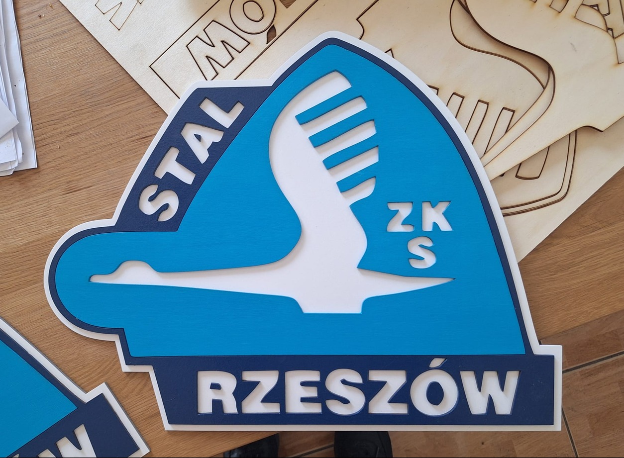 Wycięty laserowo, warstwowy, niebiesko-biały herb 'Stal Rzeszów ZKS' z drewna. Widoczne elementy wycięte z surowego drewna w tle.