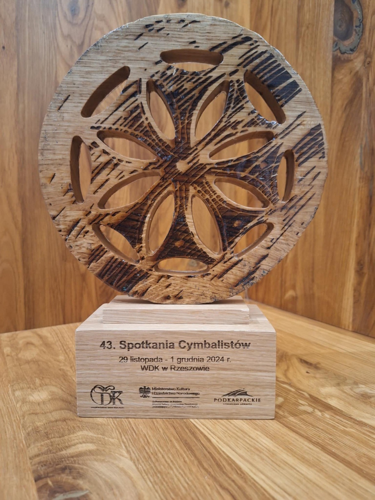 Drewniana statuetka z ażurowym wzorem kwiatowym, na podstawie z napisem '43. Spotkania Cymbalistów', na tle drewnianej ściany. Unikatowy design i naturalne materiały.