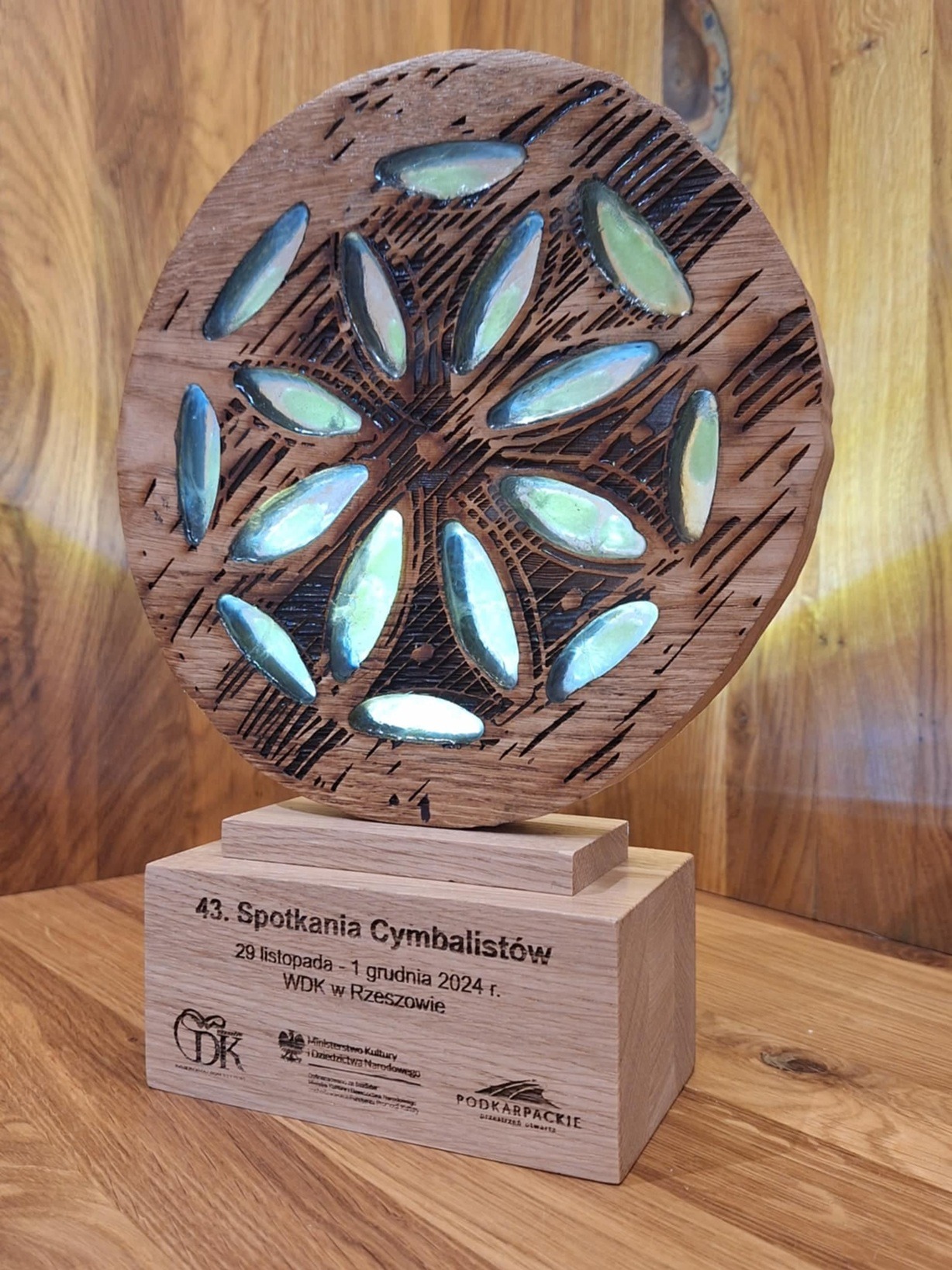 Drewniana nagroda z wygrawerowanym wzorem kwiatowym i datą wydarzenia, stojąca na drewnianej podstawie z napisem '43. Spotkania Cymbalistów'.