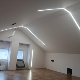 Patryk Januszewski - Oświetlenie LED w skosach poddasza, z liniowymi lampami na ścianach i suficie, tworzącymi nowoczesny design. Widoczne okna i drewniana podłoga.