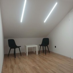 Patryk Januszewski - Minimalistyczne wnętrze poddasza z dwiema podłużnymi lampami LED na suficie, dwoma czarnymi krzesłami i białym stolikiem na drewnianej podłodze.