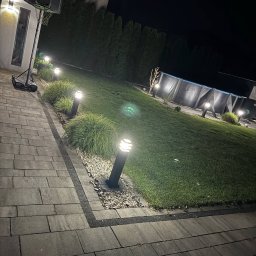 Patryk Januszewski - Oświetlony ogród nocą z lampami stojącymi wzdłuż ścieżki i podświetlonym basenem w tle, kosz do koszykówki przy budynku. Oświetlenie elewacji.