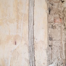 Patryk Januszewski - Białe przewody elektryczne prowadzone w bruździe, zabezpieczone uchwytami, wchodzą do pomarańczowych peszli w ścianie. Widoczne surowe wykończenie ściany i metalowy profil sufitowy.