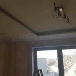 Sufit podwieszany z widocznymi przewodami elektrycznymi i zamontowanym oświetleniem. W tle okno z widokiem na zabudowania.