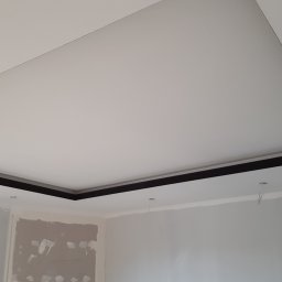 Patryk Januszewski - Podwieszany sufit z czarną listwą w wykończonym wnętrzu. Widoczne przygotowania do montażu oświetlenia, z wystającymi przewodami. Ściany w trakcie malowania.