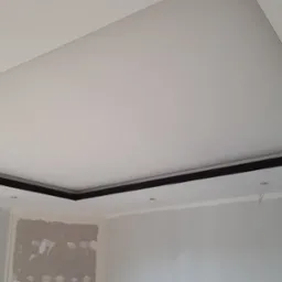 Podwieszany sufit z czarną listwą w wykończonym wnętrzu. Widoczne przygotowania do montażu oświetlenia, z wystającymi przewodami. Ściany w trakcie malowania.