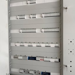 Patryk Januszewski - Otwarta, szara rozdzielnica elektryczna z wieloma rzędami białych wyłączników nadprądowych i różnicowoprądowych, niektóre z numeracją, zamontowana na surowej ścianie. Klucz wisi na drzwiach.