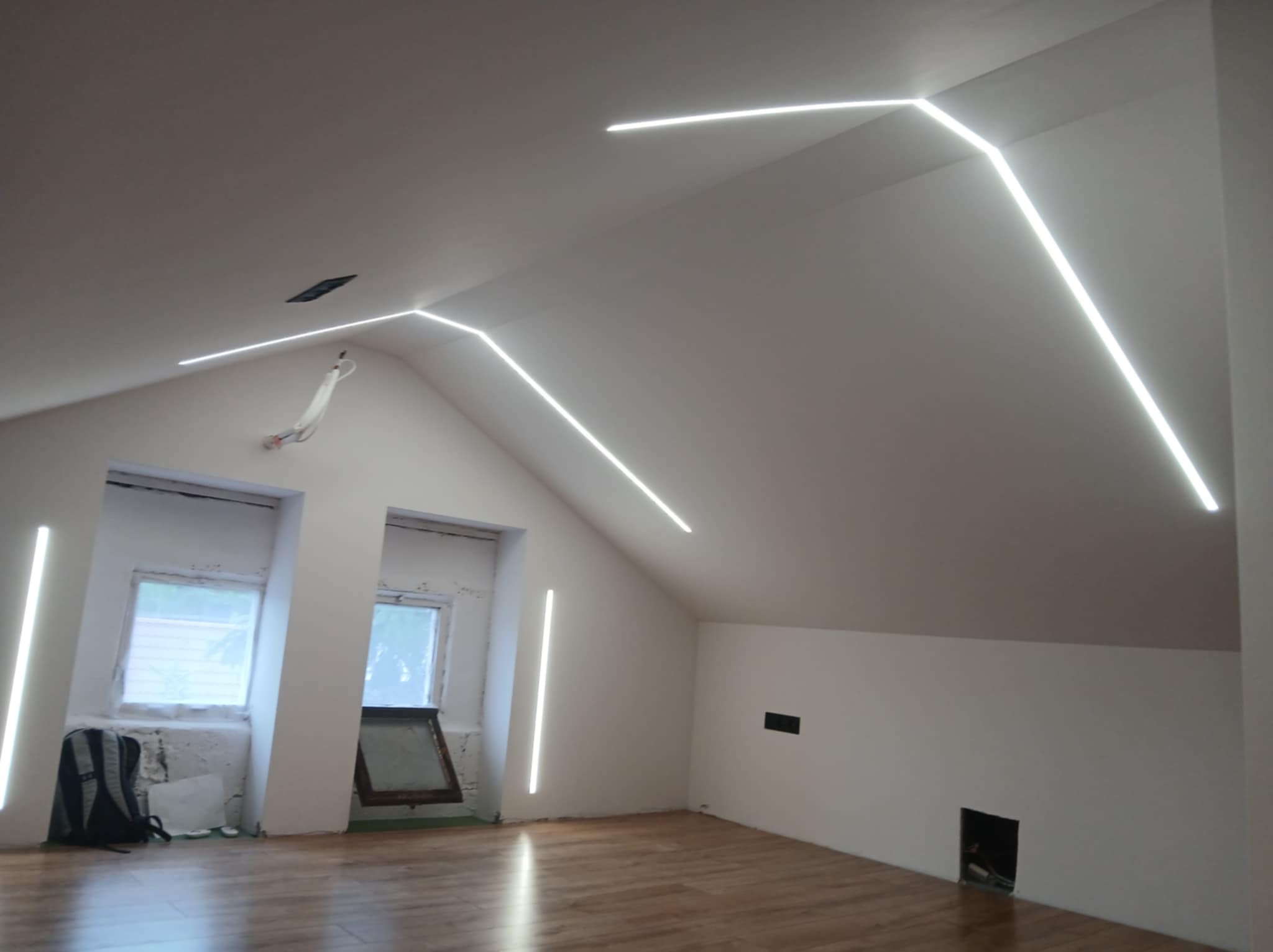 Oświetlenie LED w skosach poddasza, z liniowymi lampami na ścianach i suficie, tworzącymi nowoczesny design. Widoczne okna i drewniana podłoga.