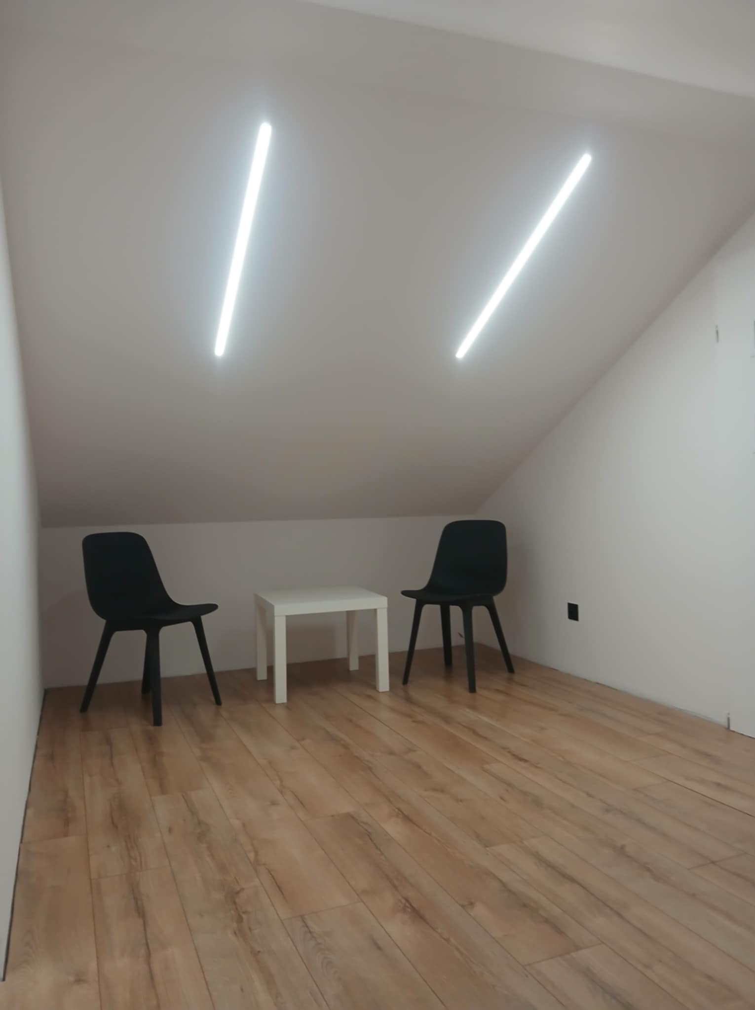 Minimalistyczne wnętrze poddasza z dwiema podłużnymi lampami LED na suficie, dwoma czarnymi krzesłami i białym stolikiem na drewnianej podłodze.