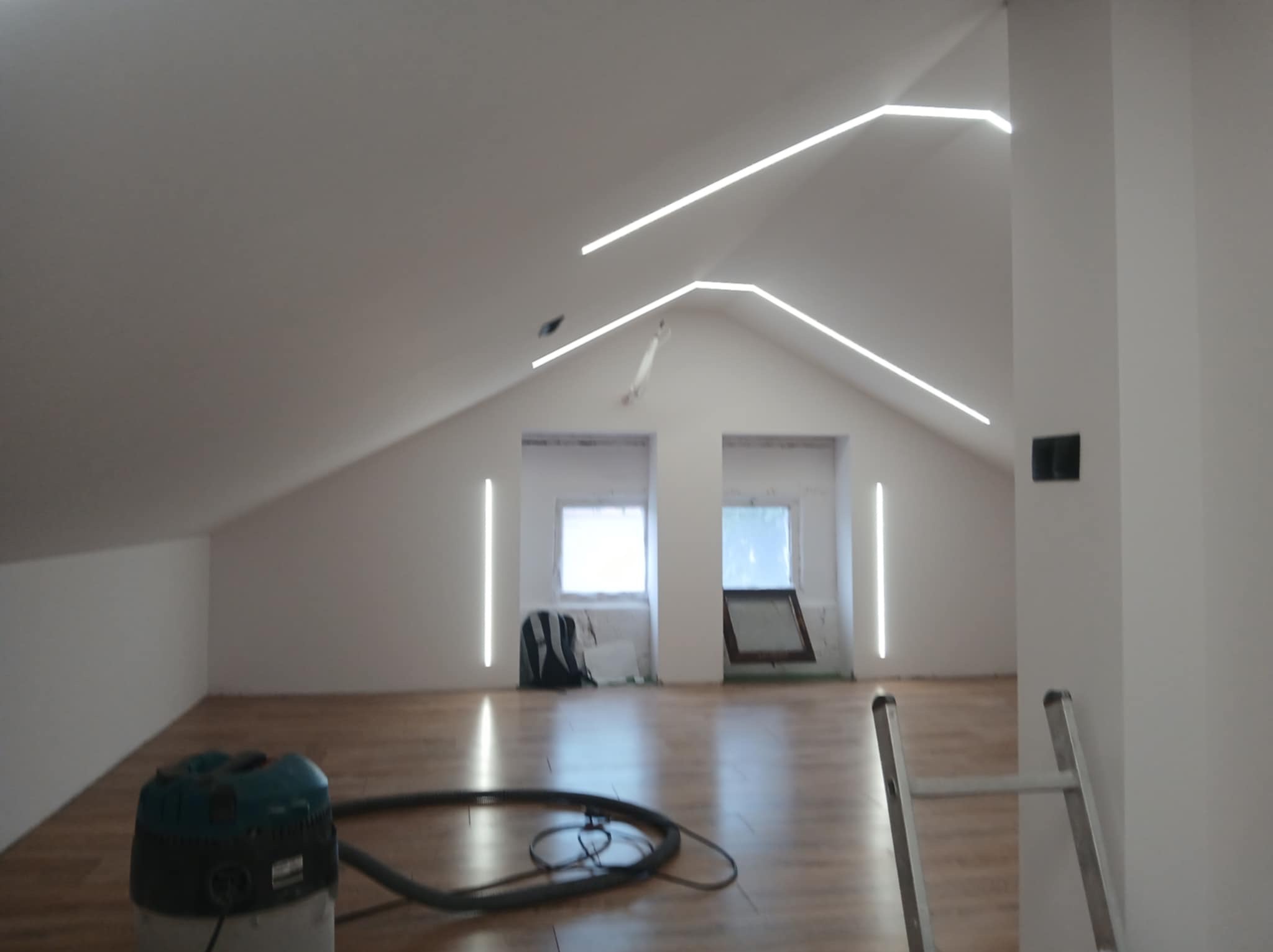 Oświetlenie LED w poddaszu: białe, liniowe lampy tworzą nowoczesny wzór na suficie. Widoczny odkurzacz i drabina, sugerujące prace wykończeniowe.
