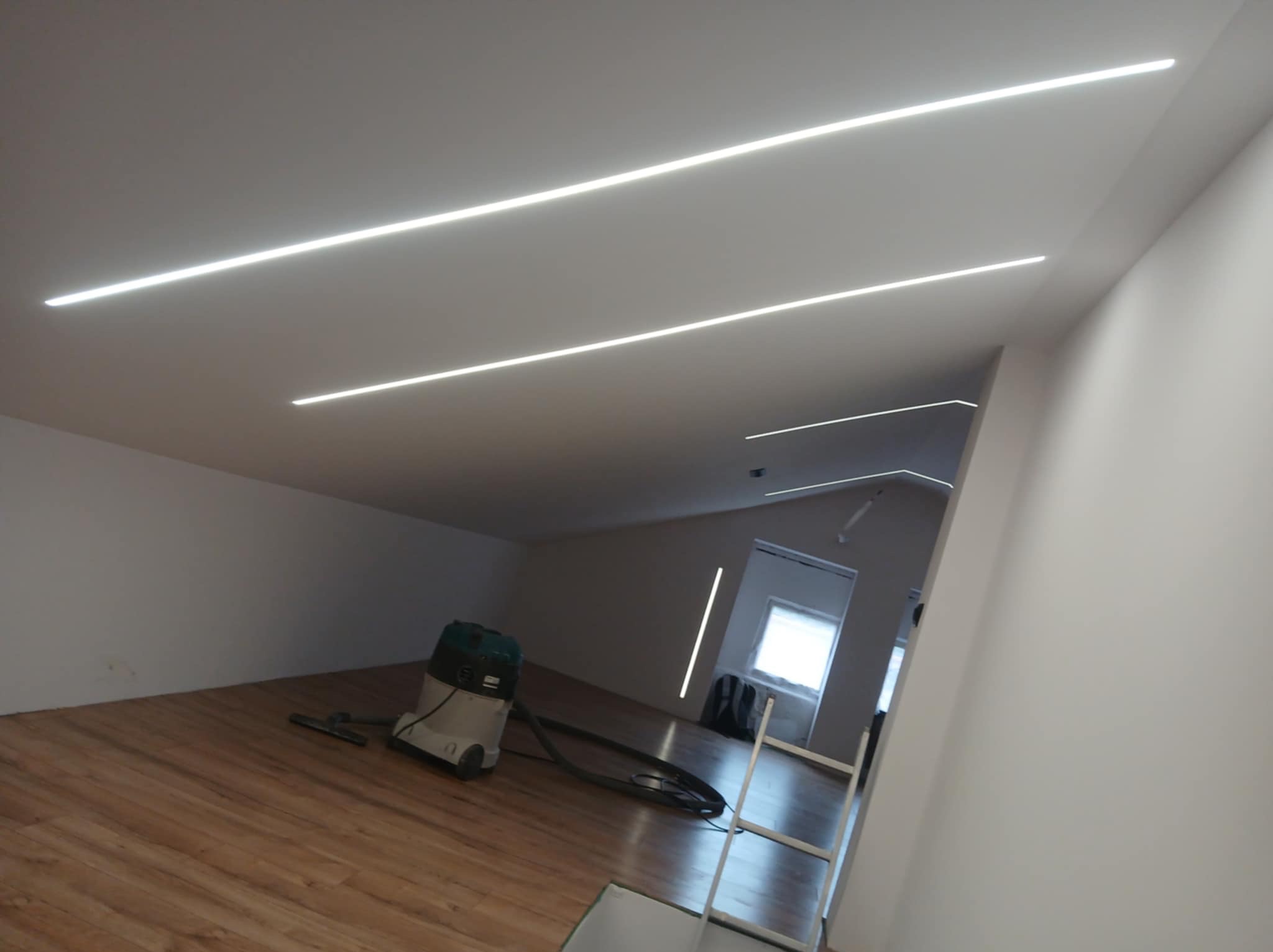 Nowoczesne oświetlenie LED liniowe zamontowane w suficie, widoczne w przestronnym wnętrzu z drewnianą podłogą, odkurzacz budowlany w rogu pomieszczenia.