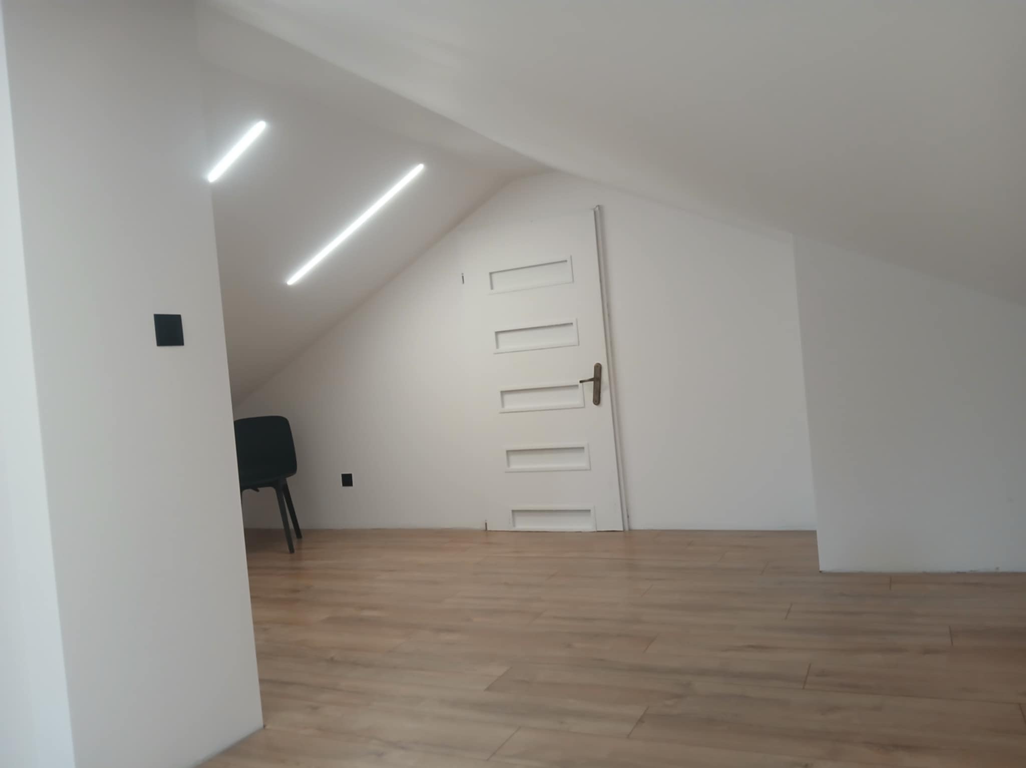 Nowoczesne oświetlenie LED w skosach poddasza, z minimalistycznym wykończeniem i drewnianą podłogą. Widoczne detale: czarny włącznik i krzesło w tle.