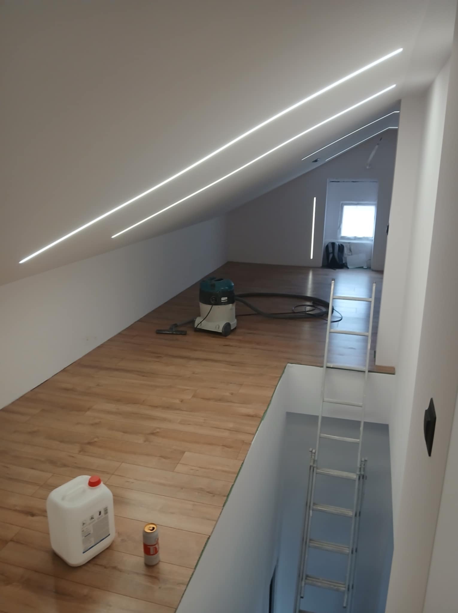 Poddasze po montażu oświetlenia liniowego LED w skosach sufitu. Widoczny odkurzacz, drabina i butelka chemii. Parkiet ułożony, gotowy do użytku.