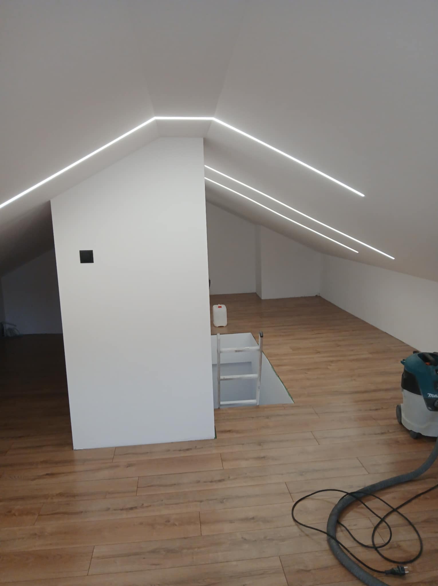 Nowoczesne oświetlenie LED w poddaszu z drewnianą podłogą. Widoczne linie świetlne w suficie i czarny włącznik na ścianie. Na podłodze odkurzacz.