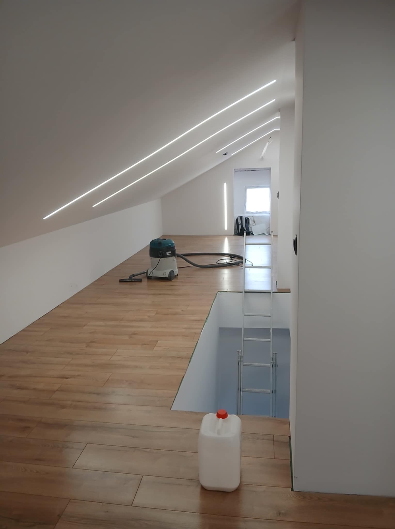 Poddasze z drewnianą podłogą i zamontowanym oświetleniem LED. Na podłodze widoczny odkurzacz i pojemnik. W tle drabina prowadząca na niższy poziom.