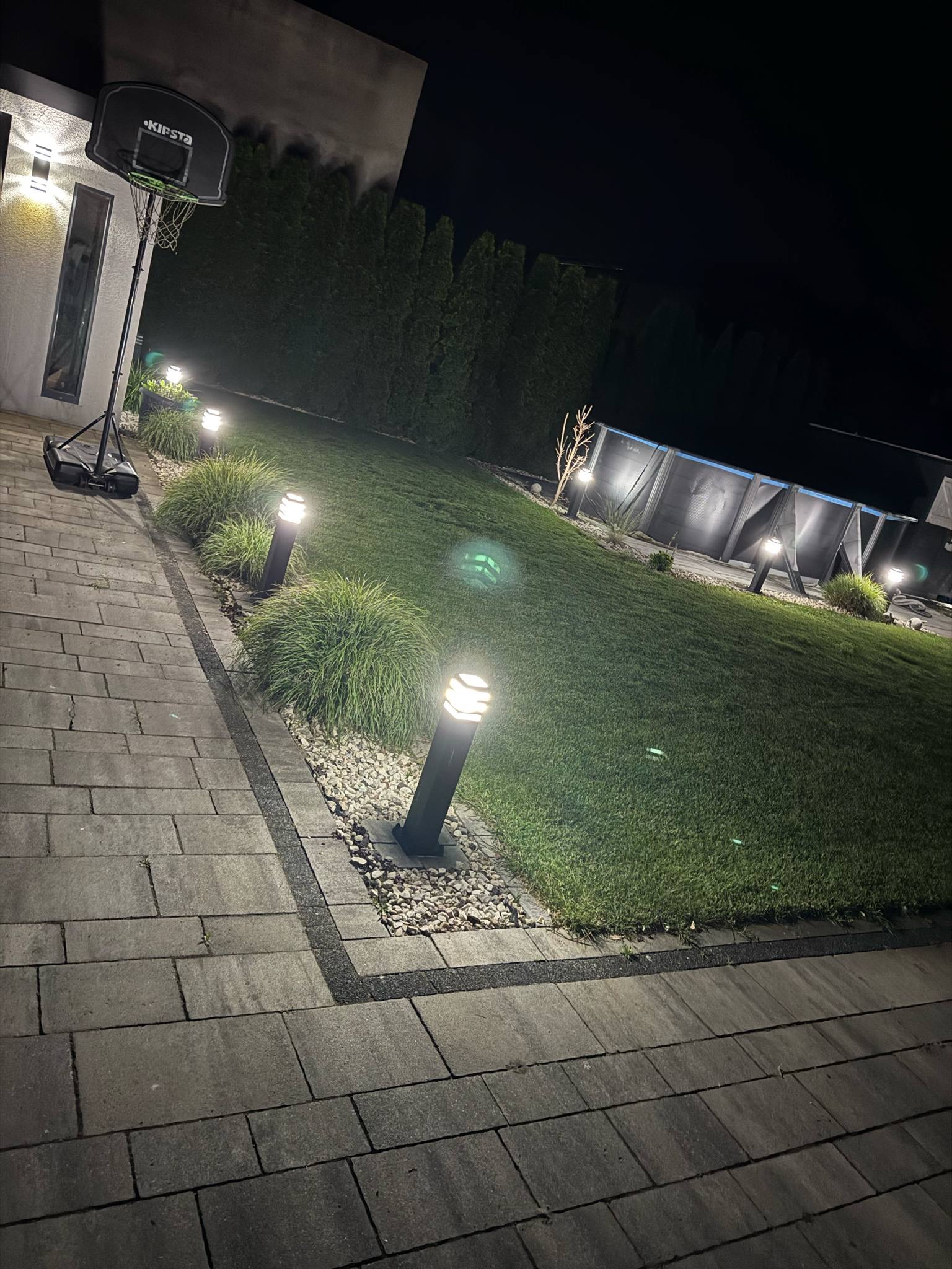 Oświetlony ogród nocą z lampami stojącymi wzdłuż ścieżki i podświetlonym basenem w tle, kosz do koszykówki przy budynku. Oświetlenie elewacji.
