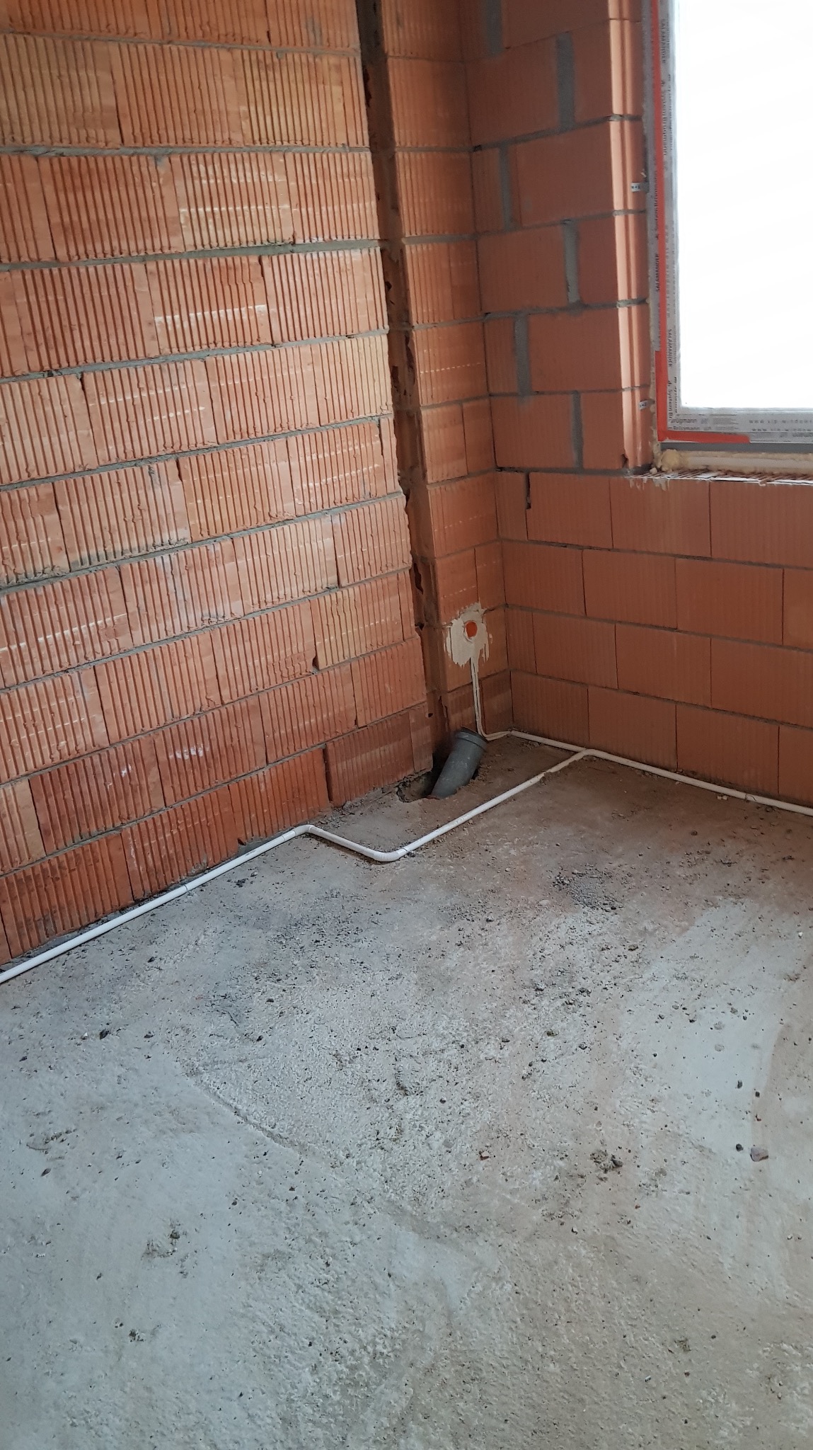 Wnętrze w trakcie budowy z widoczną instalacją elektryczną w białej rurce PCV prowadzoną po podłodze i wzdłuż ściany z cegły. Widoczny otwór w ścianie i okno.