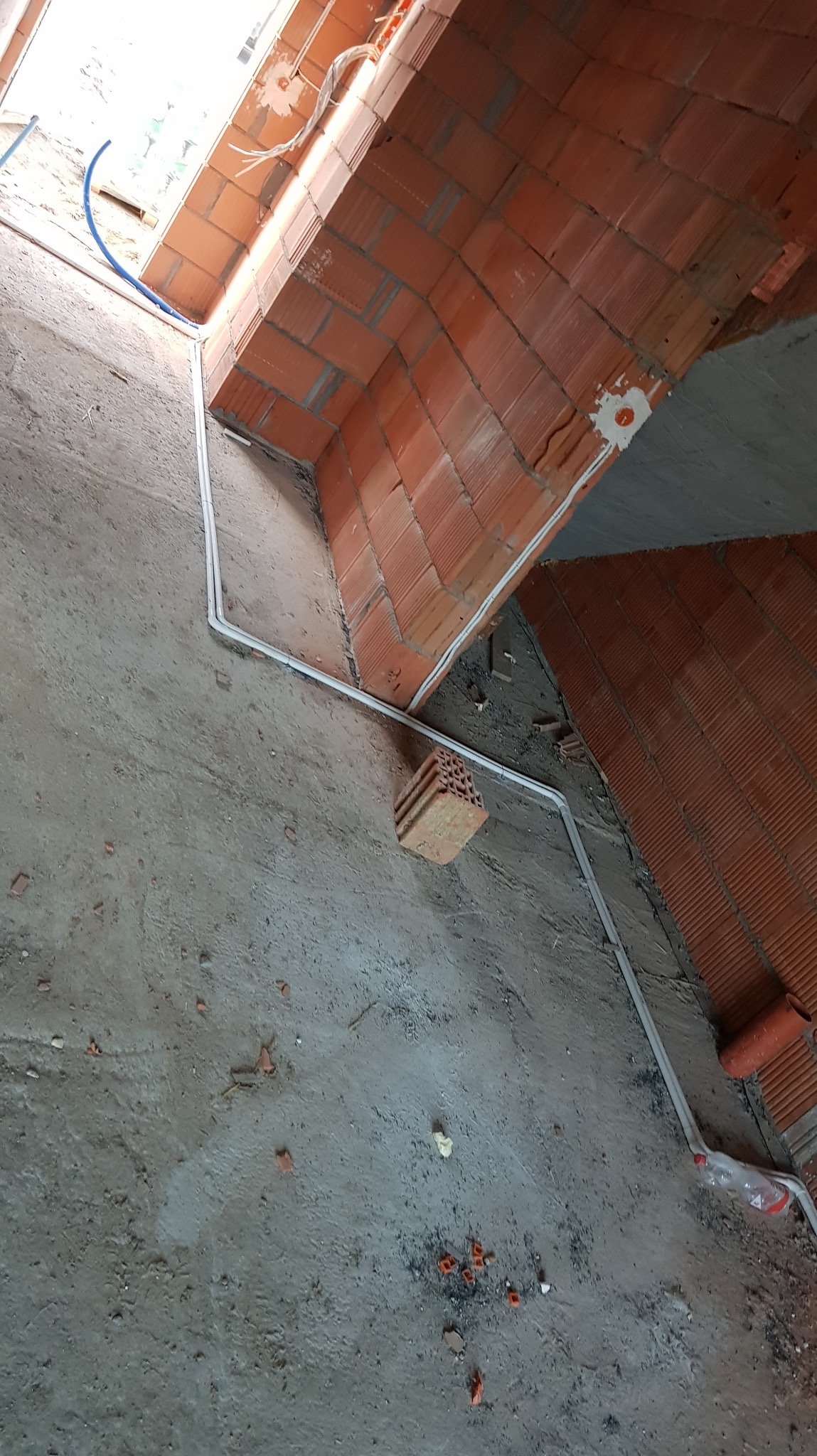 Wnętrze w trakcie budowy z widocznymi białymi rurkami instalacyjnymi poprowadzonymi wzdłuż ścian z cegły dziurawki i surową podłogą. Widok z góry.