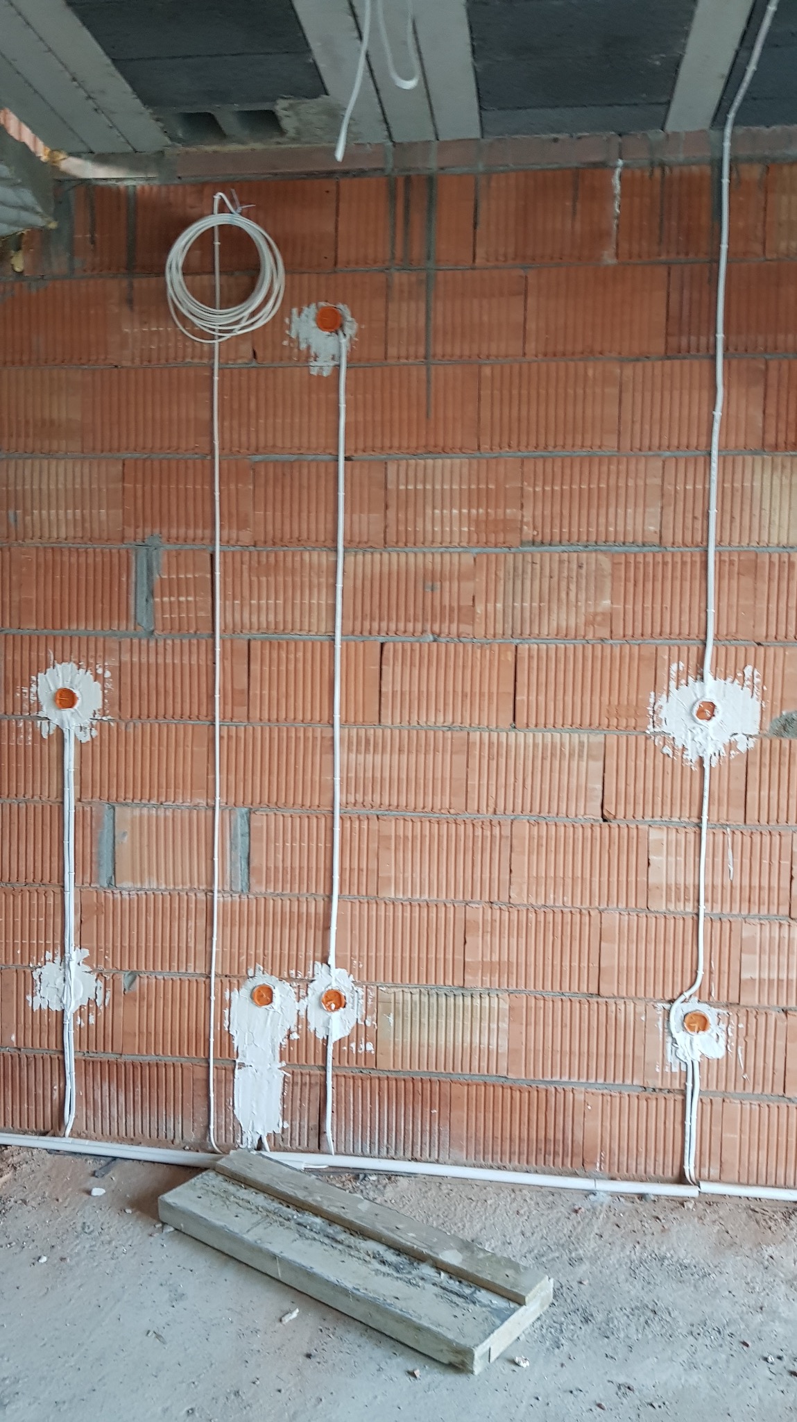 Instalacja elektryczna w trakcie montażu na ceglanej ścianie. Widoczne kable w białych rurkach, puszki elektryczne zalane gipsem, surowy stan wykończenia.