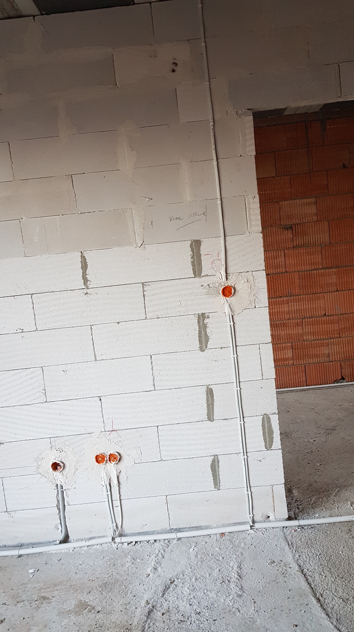 Biała ściana z bloków z widocznymi puszkami i przewodami elektrycznymi w trakcie montażu, surowy stan wykończenia wnętrza. Widoczne ślady zaprawy.