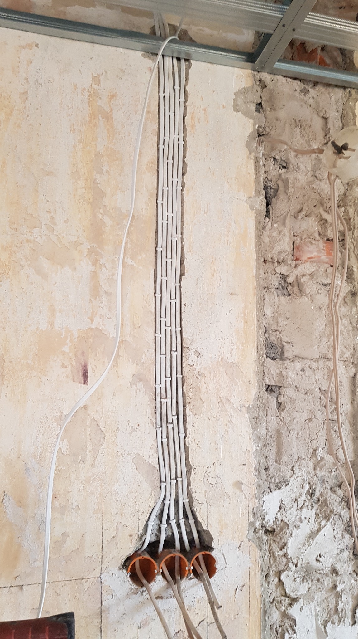 Białe przewody elektryczne prowadzone w bruździe, zabezpieczone uchwytami, wchodzą do pomarańczowych peszli w ścianie. Widoczne surowe wykończenie ściany i metalowy profil sufitowy.