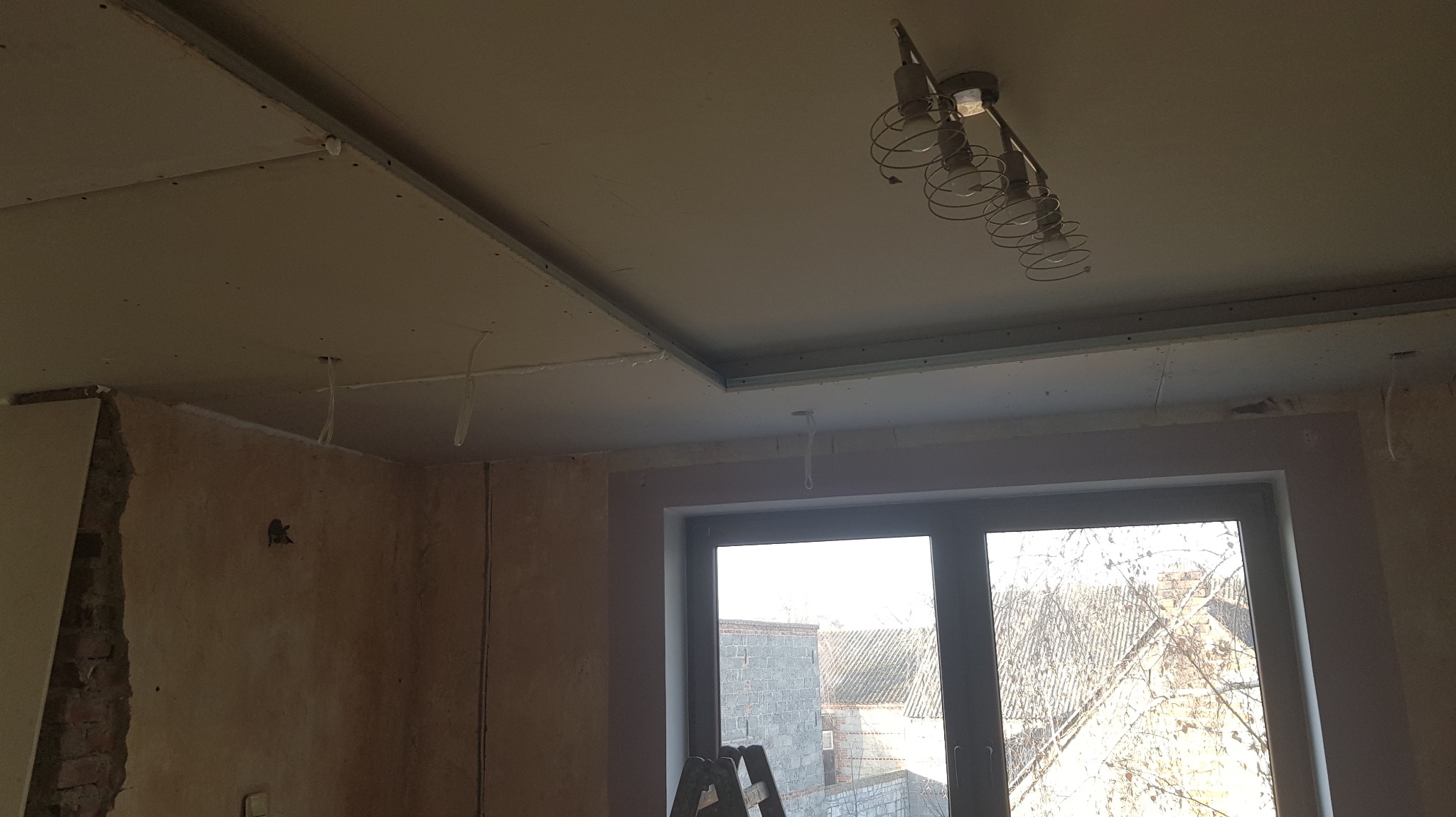 Sufit podwieszany z widocznymi przewodami elektrycznymi i zamontowanym oświetleniem. W tle okno z widokiem na zabudowania.