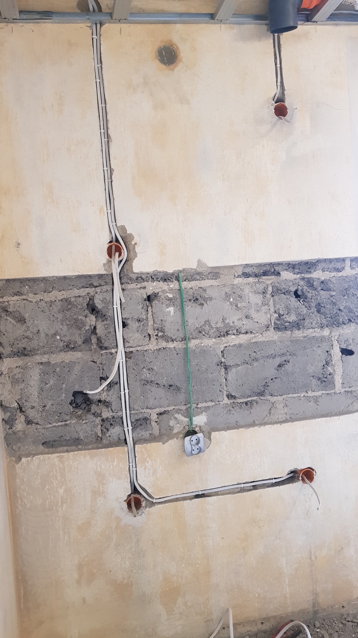 Ściana w trakcie remontu z widocznymi przewodami elektrycznymi w peszlach, puszki instalacyjne i gniazdko. Fragment sufitu podwieszanego w tle.