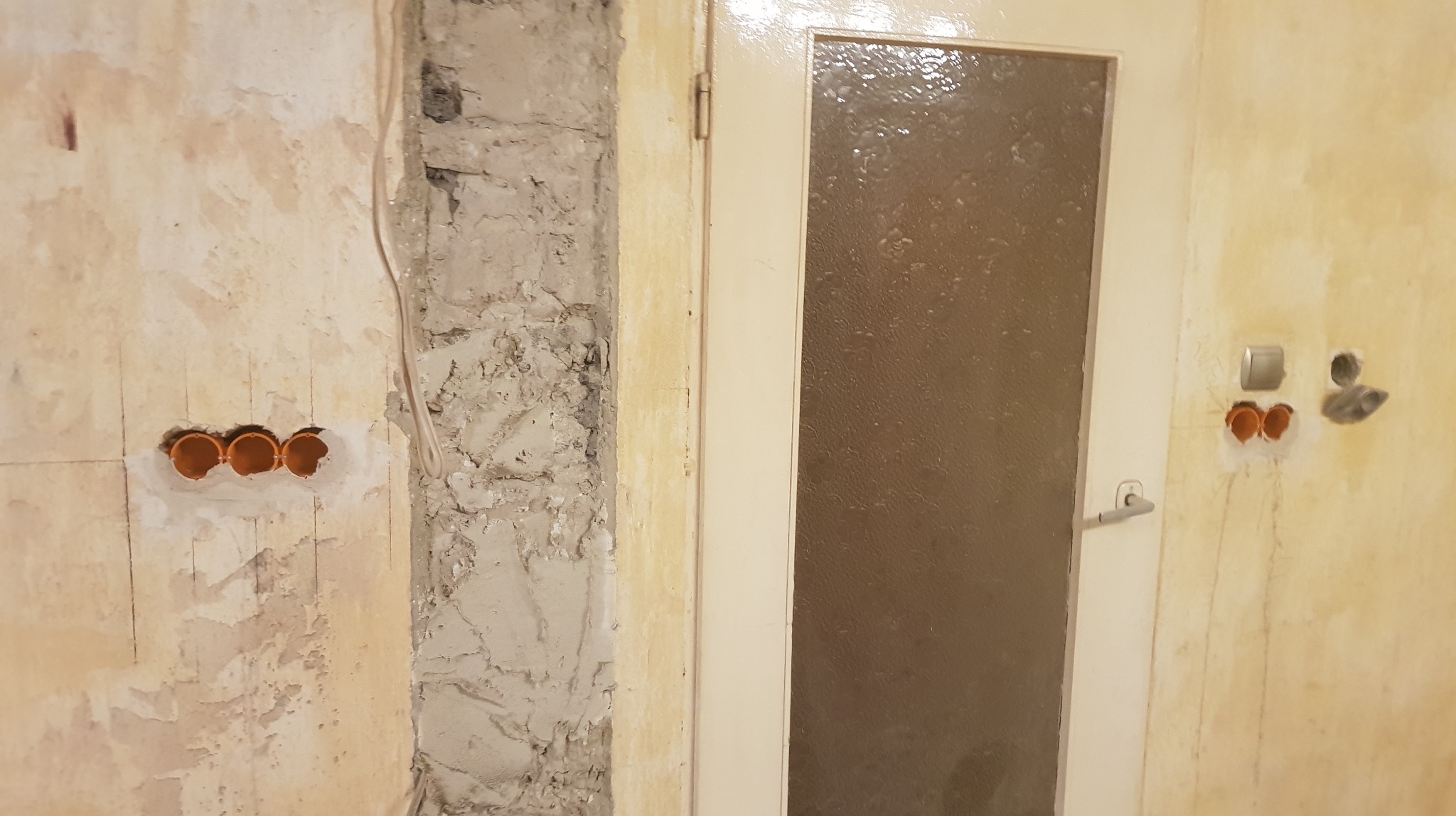 Ściana z trzema pomarańczowymi puszkami instalacyjnymi, obok drzwi z szybą. Widoczne ślady po kuciu i resztki starej farby na ścianie. Przewód elektryczny wzdłuż ściany.
