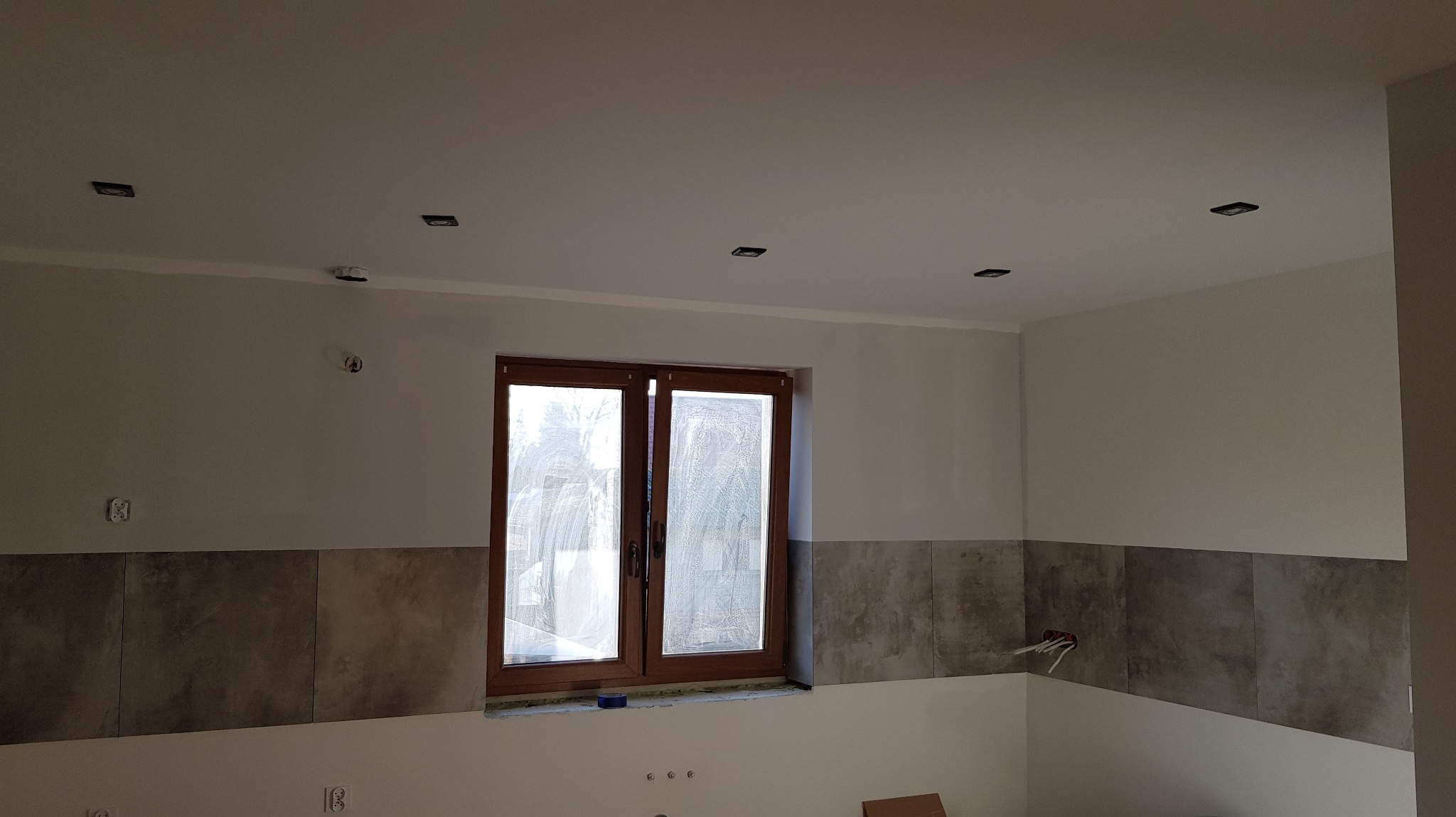 Wnętrze w trakcie remontu: zamontowane płytki ścienne, okno z brązową ramą i częściowo zamontowane, kwadratowe lampy sufitowe w nowym mieszkaniu.