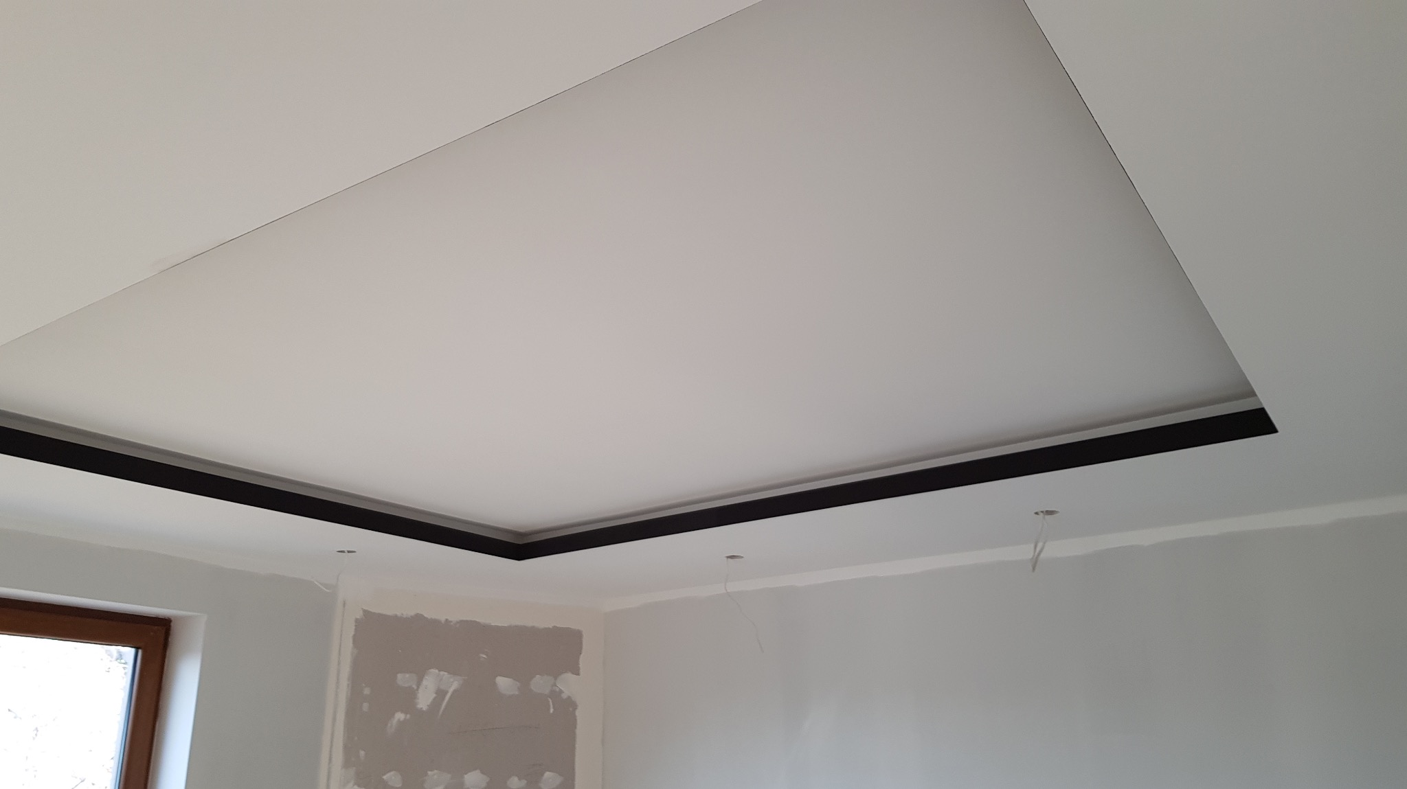 Podwieszany sufit z czarną listwą w wykończonym wnętrzu. Widoczne przygotowania do montażu oświetlenia, z wystającymi przewodami. Ściany w trakcie malowania.