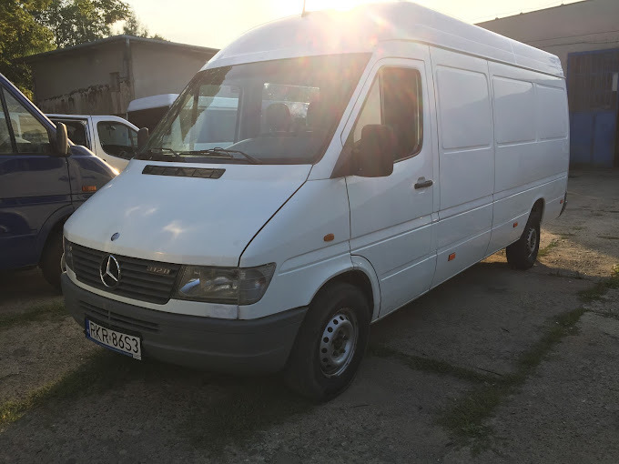 Biały Mercedes Sprinter 312D z widoczną tablicą rejestracyjną PKR 86S3, zaparkowany na zewnątrz, z delikatnym blaskiem słońca odbijającym się od karoserii.