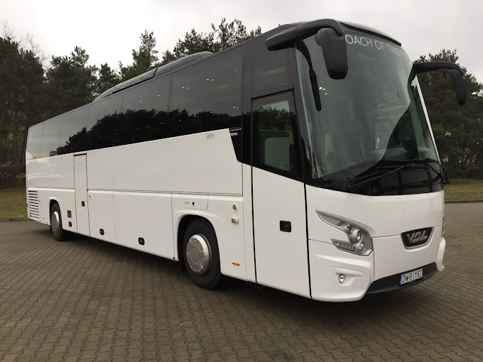 Nowoczesny, biały autokar VDL z czarnymi szybami i napisem 'COACH OF THE YEAR' nad przednią szybą, zaparkowany na tle drzew.
