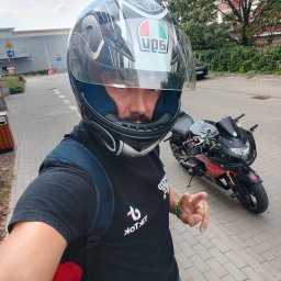 EKOSPÓŁKA Sp. z o. o. - Mężczyzna w kasku motocyklowym, z plecakiem, pozuje na tle czarno-czerwonego motocykla na kostce brukowej, z budynkiem w tle. Kask z logo. Ujęcie z dołu.