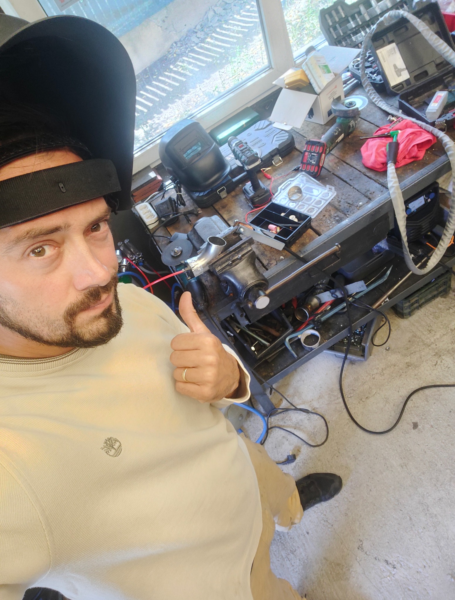 Autoportret spawacza w masce, z kciukiem w górę, na tle warsztatu z narzędziami i fragmentem spawanej rury w imadle.