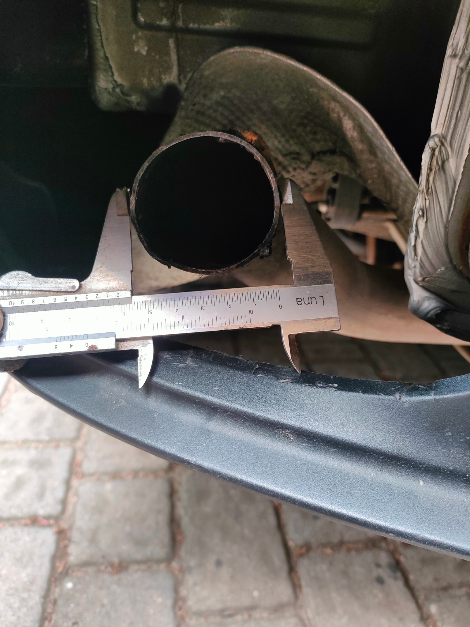 Pomiar średnicy rury wydechowej podwozia samochodu za pomocą suwmiarki Luna. Widoczne fragmenty podwozia i kostki brukowej. Pomiar precyzyjny.