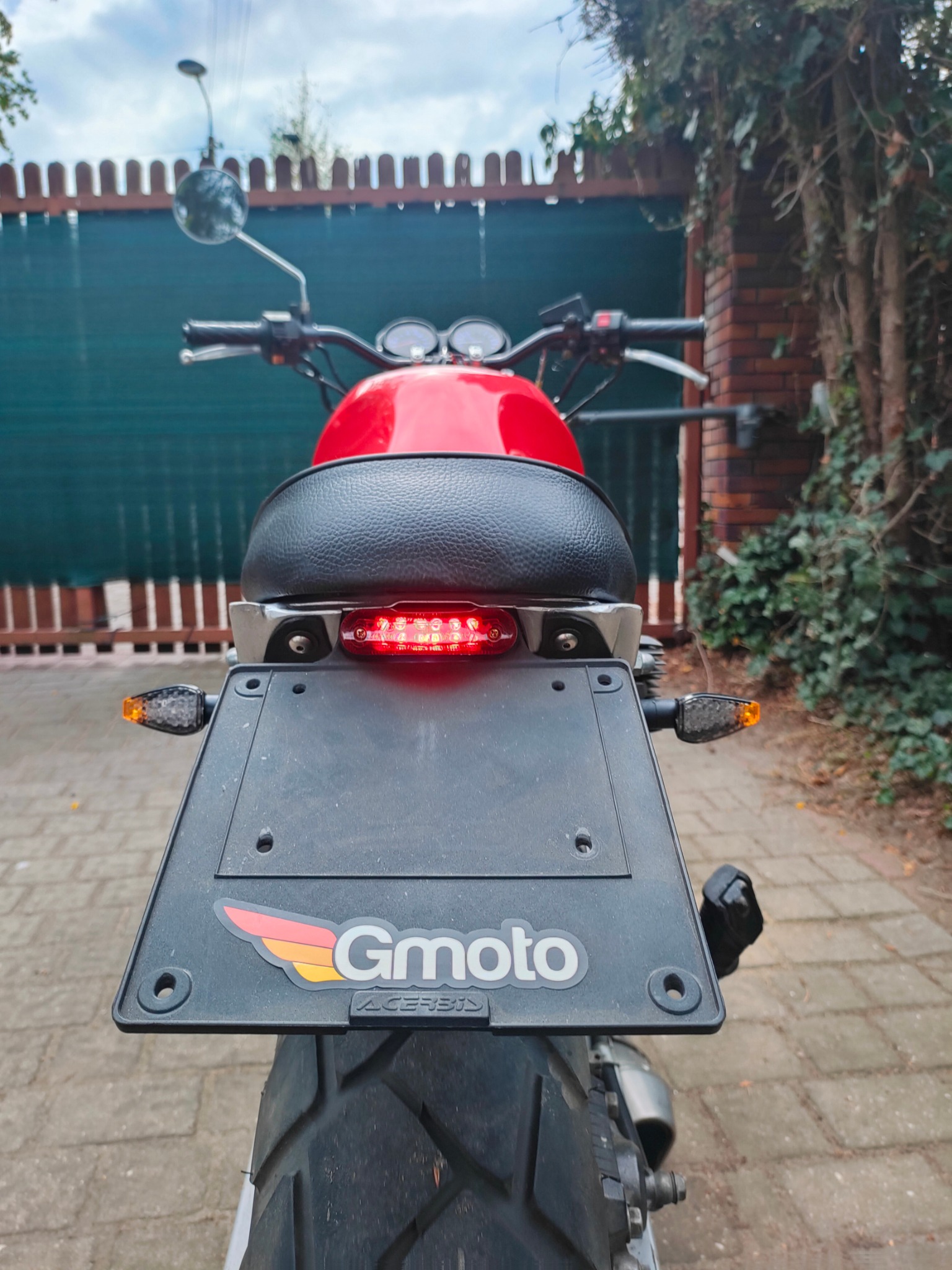 Tył motocykla Gmoto z czerwonym bakiem i czarnym siedzeniem na tle zielonego ogrodzenia i ceglanego muru porośniętego bluszczem. Widoczny błotnik i oświetlenie.