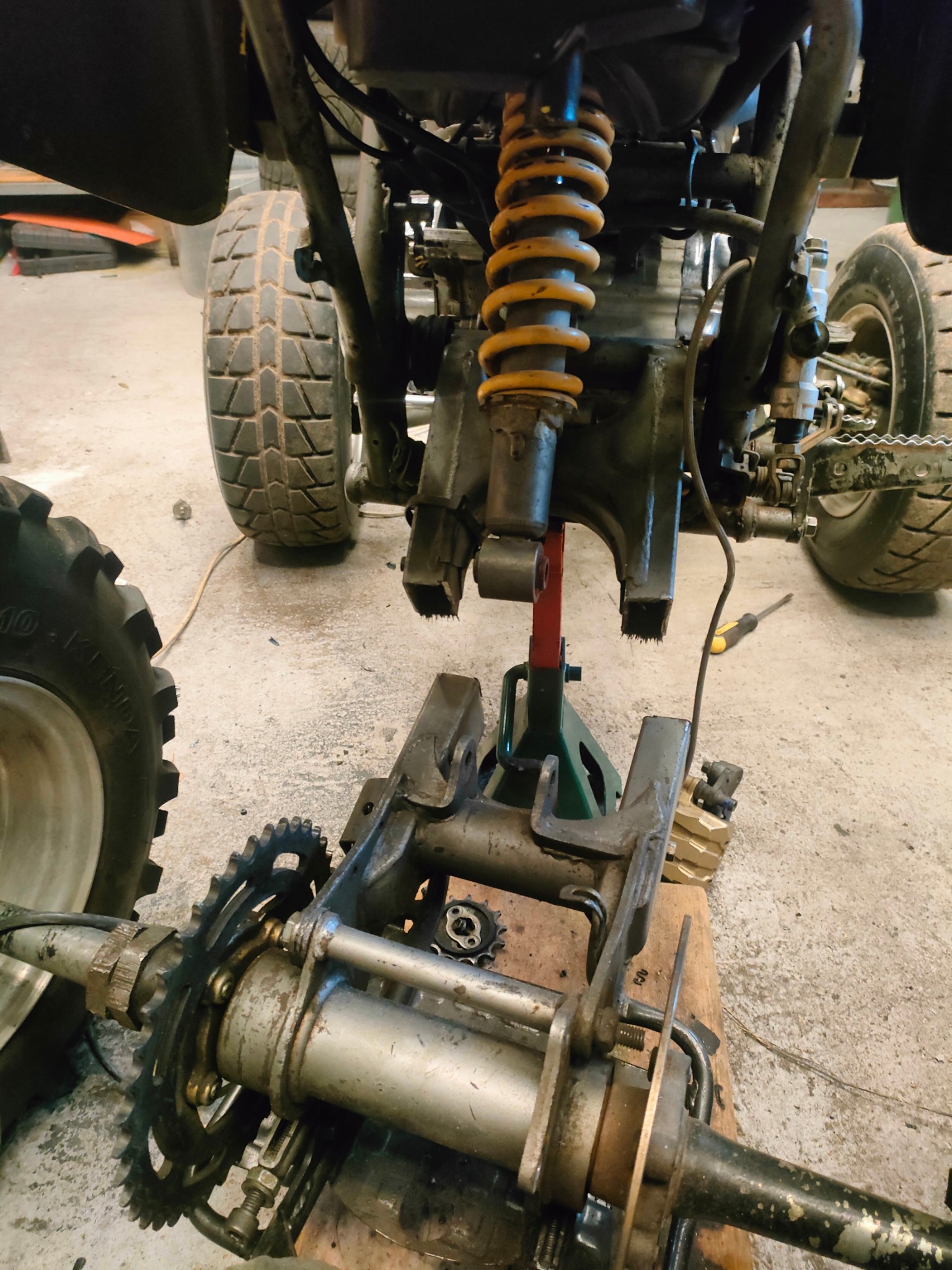 Demontaż tylnego zawieszenia quada z widocznym amortyzatorem, łańcuchem i zębatką. Elementy wsparte na podnośniku warsztatowym.