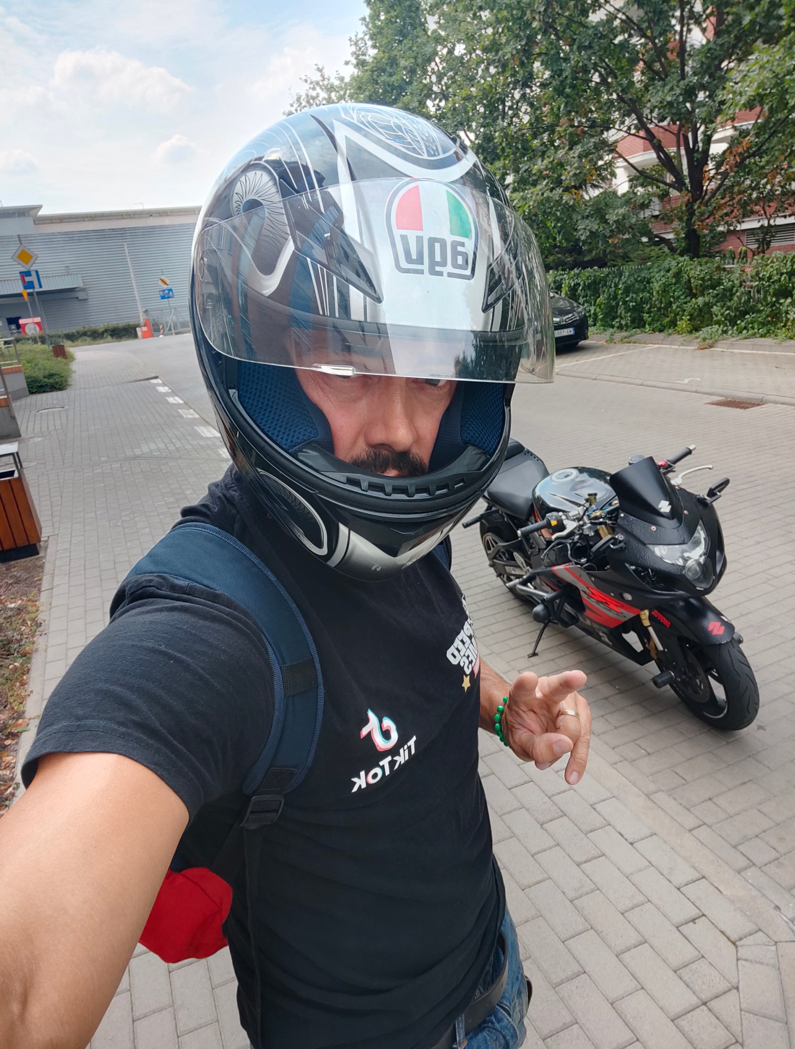 Mężczyzna w kasku motocyklowym, z plecakiem, pozuje na tle czarno-czerwonego motocykla na kostce brukowej, z budynkiem w tle. Kask z logo. Ujęcie z dołu.
