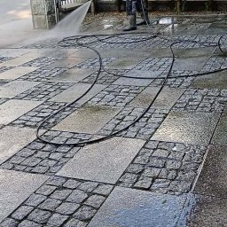 ZIELONA EKIPA KRZYSZTOF ŁABA - Czyszczenie kostki brukowej myjką ciśnieniową. Mężczyzna w roboczym stroju czyści kamienną kostkę brukową na posesji. Widoczny wąż i lanca myjki.