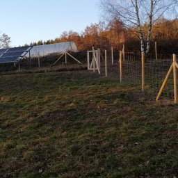 ZIELONA EKIPA KRZYSZTOF ŁABA - Siatka ogrodzeniowa z drewnianymi słupkami na nierównym terenie, w tle panele słoneczne i tunel foliowy. Widok na wzgórze o zmierzchu.