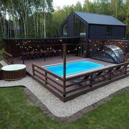 KM DESIGN SPA KRZYSZTOF KACZMARSKI - Nowoczesny taras z basenem i jacuzzi, otoczony drewnianą zabudową i dekoracyjnym oświetleniem. W tle dom z ciemną elewacją i las.