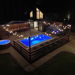 KM DESIGN SPA KRZYSZTOF KACZMARSKI - Oświetlony basen i jacuzzi na drewnianym tarasie nocą. W tle budynek z oknami i zadaszona jadalnia. Dekoracyjne lampki dodają uroku przestrzeni.