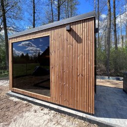 KM DESIGN SPA KRZYSZTOF KACZMARSKI - Nowoczesna sauna ogrodowa z drewnianą elewacją i dużym oknem, odbijającym błękitne niebo. Obok jacuzzi na wyłożonym kostką tarasie, otoczona zielenią.