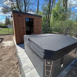 KM DESIGN SPA KRZYSZTOF KACZMARSKI - Nowoczesna sauna z drewna obok czarnego jacuzzi z pokrywą, na tle zieleni i błękitnego nieba. Widoczne prace wykończeniowe wokół.