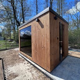 KM DESIGN SPA KRZYSZTOF KACZMARSKI - Nowoczesna sauna zewnętrzna z drewna, z dużym oknem odbijającym błękitne niebo. Minimalistyczny design, otoczona zielenią i elementami przygotowanego terenu.