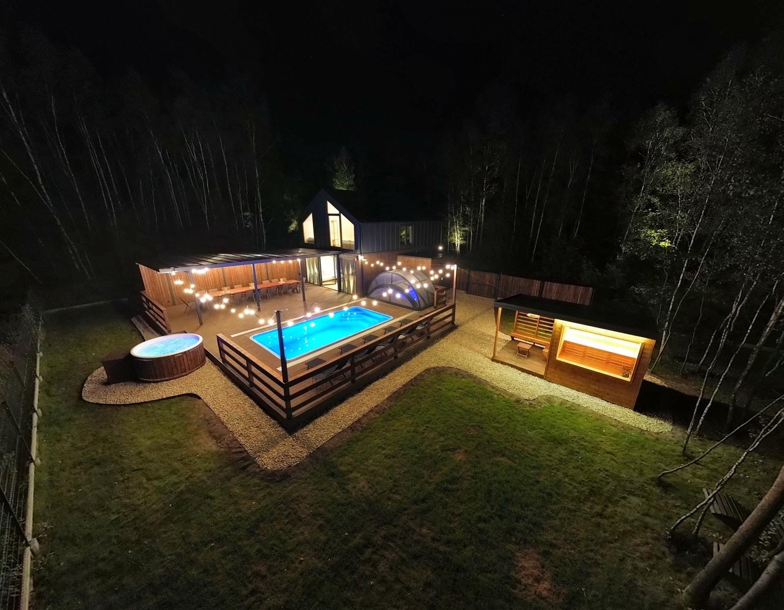 Oświetlony basen z tarasem, jacuzzi i sauną w ogrodzie nocą. Drewniana architektura, dekoracyjne lampki, ciemne tło z drzewami.