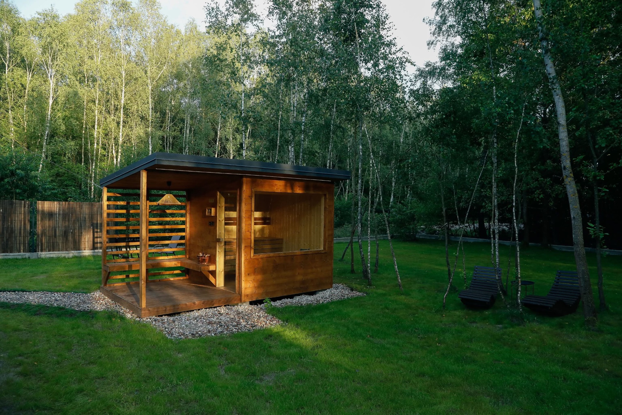 Drewniana sauna ogrodowa z przeszkloną ścianą, zadaszonym tarasem i ażurowymi ściankami, otoczona zielonym trawnikiem i brzozowym lasem. Dwa leżaki w tle.