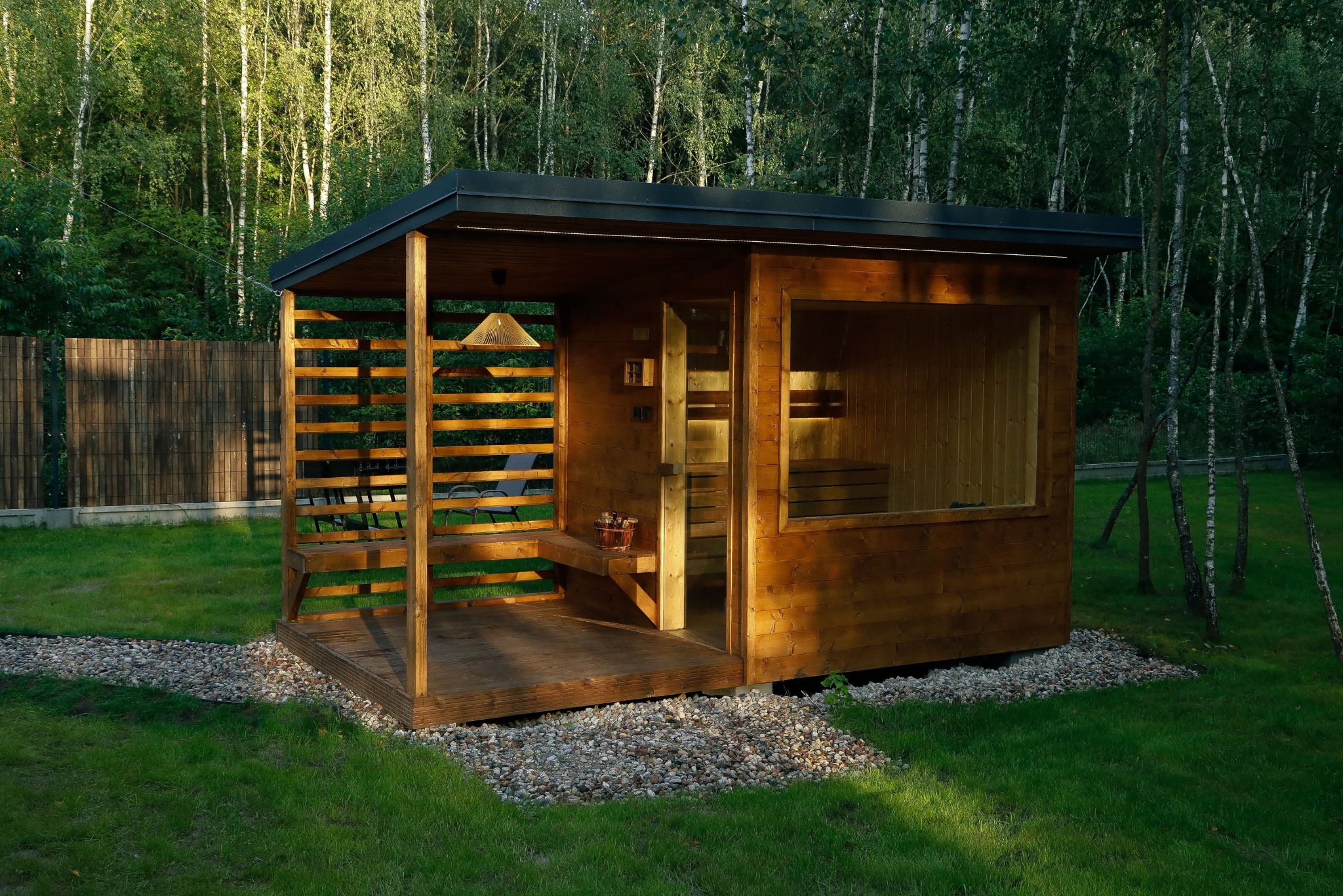 Drewniana sauna z zadaszeniem i ażurową ścianą na trawniku, oświetlona wiszącą lampą. Kamienna ścieżka prowadzi do wejścia. W tle drzewa.